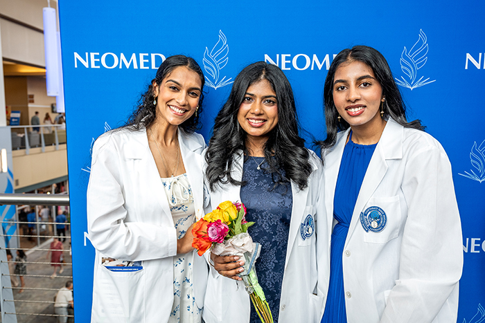 three young women in medical white coats pose in front of a blue NEOMED backdrop. the woman in the center holds a bouquet of flowers