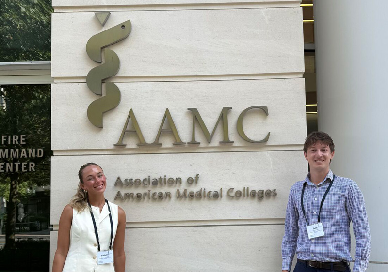 A woman in a white shirt and man in a blue shirt pose in front of the AAMC Association of American Medical Colleges sign