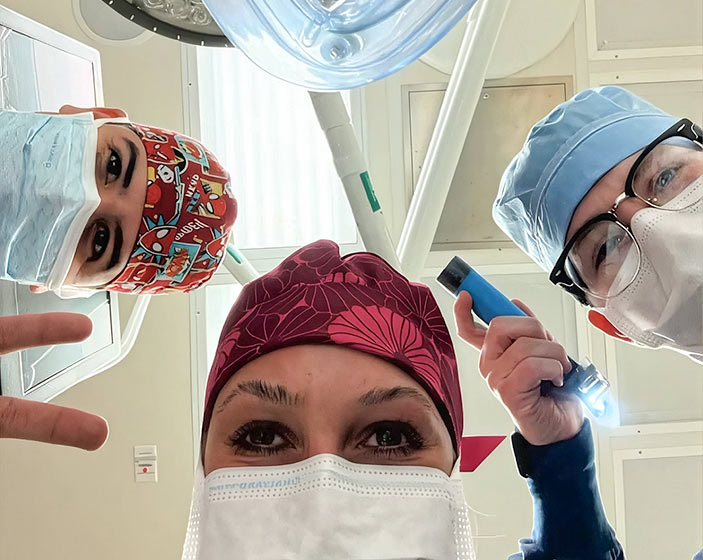 Three people wearing surgical caps and face masks look down toward the camera under operating room lights.