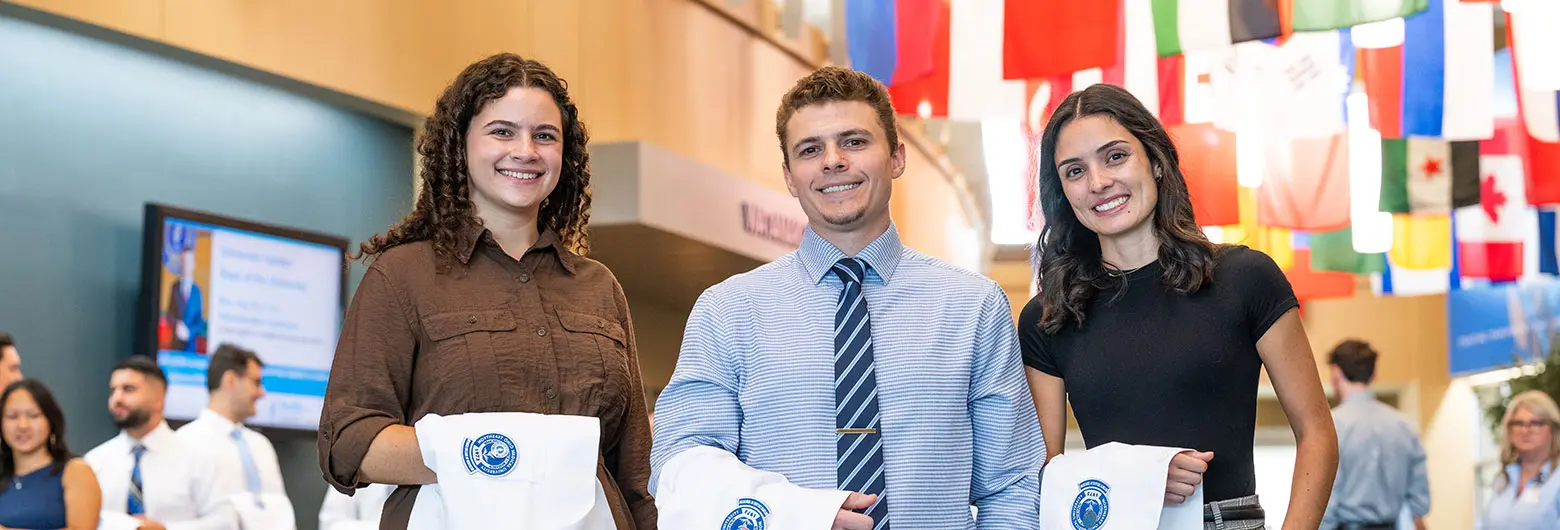 Three students smile and hold white coats in a university atrium decorated with international flags.