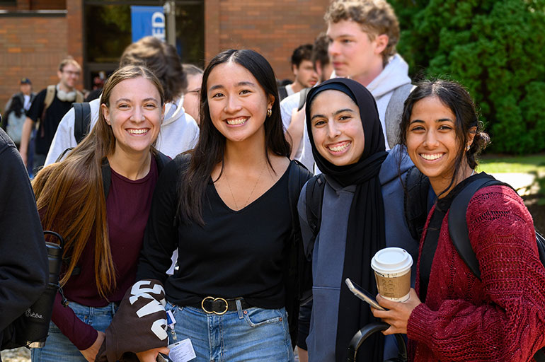 Four students smile together outdoors on a campus walkway, with other people and brick buildings in the background.