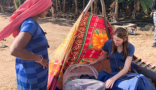 A medical student seated outdoors examines a covered infant carrier beside another person standing near a colorful fabric shelter.