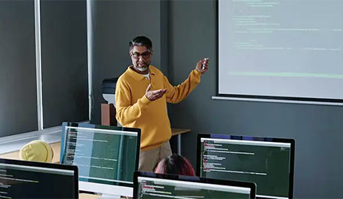A professor gestures while talking before a screen during a class.