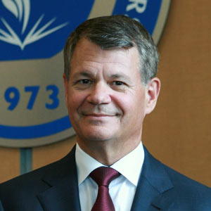A man in a dark suit and red tie poses in front of the NEOMED seal