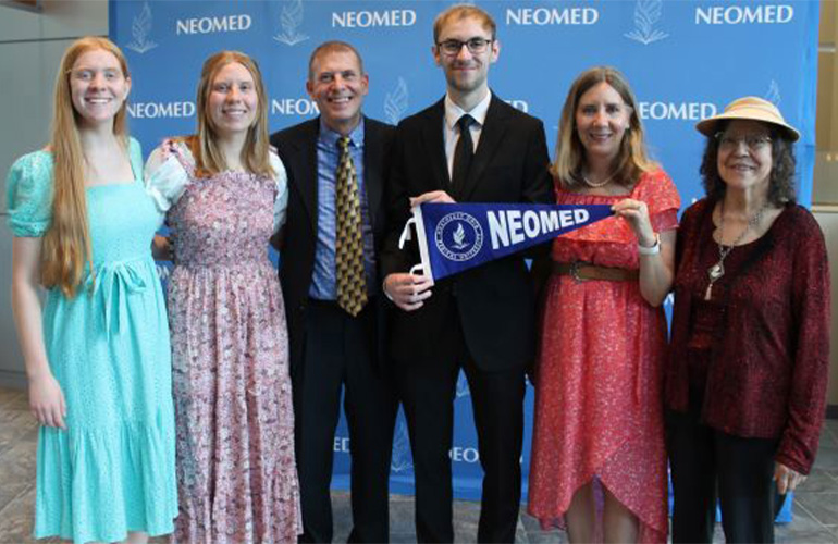 Family of six poses in front of a NEOMED step-and-repeat backdrop holding a NEOMED flag