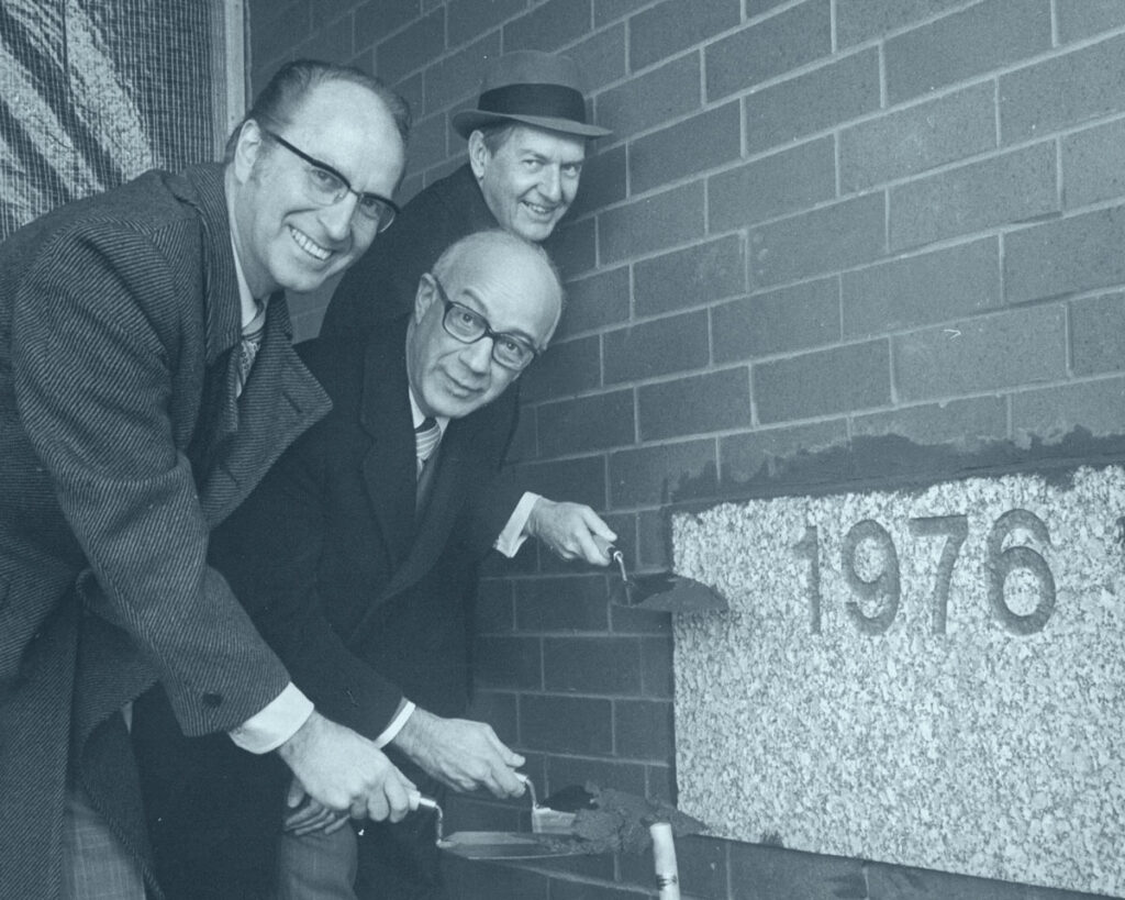 Three men in coats and ties apply mortar to a block that reads "1976."