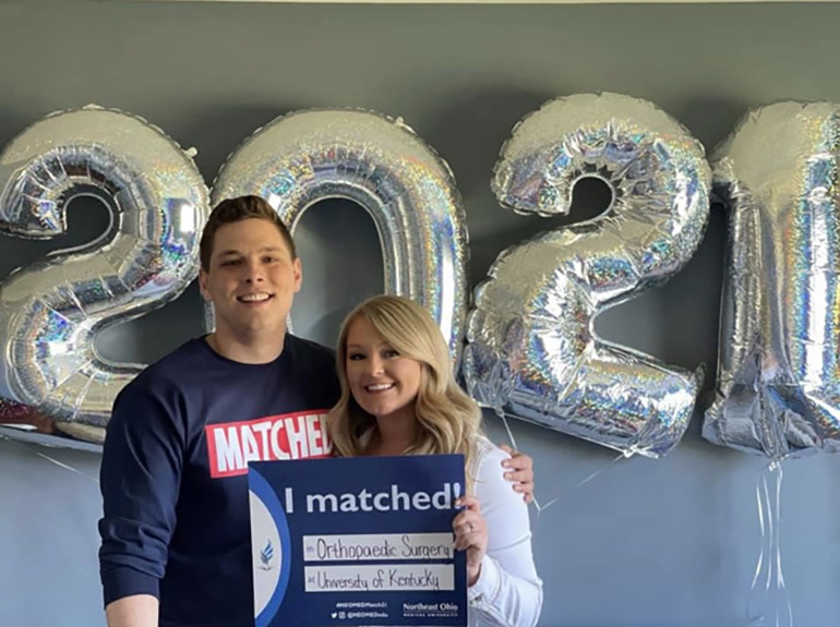 a man and a woman stand in front of silver balloons shaped like the year 2021. The woman holds a small sign that says "I matched"