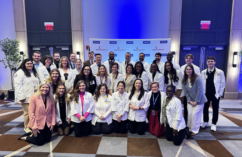 large group of people, most wearing white coats, pose in front of a step-and-repeat backdrop