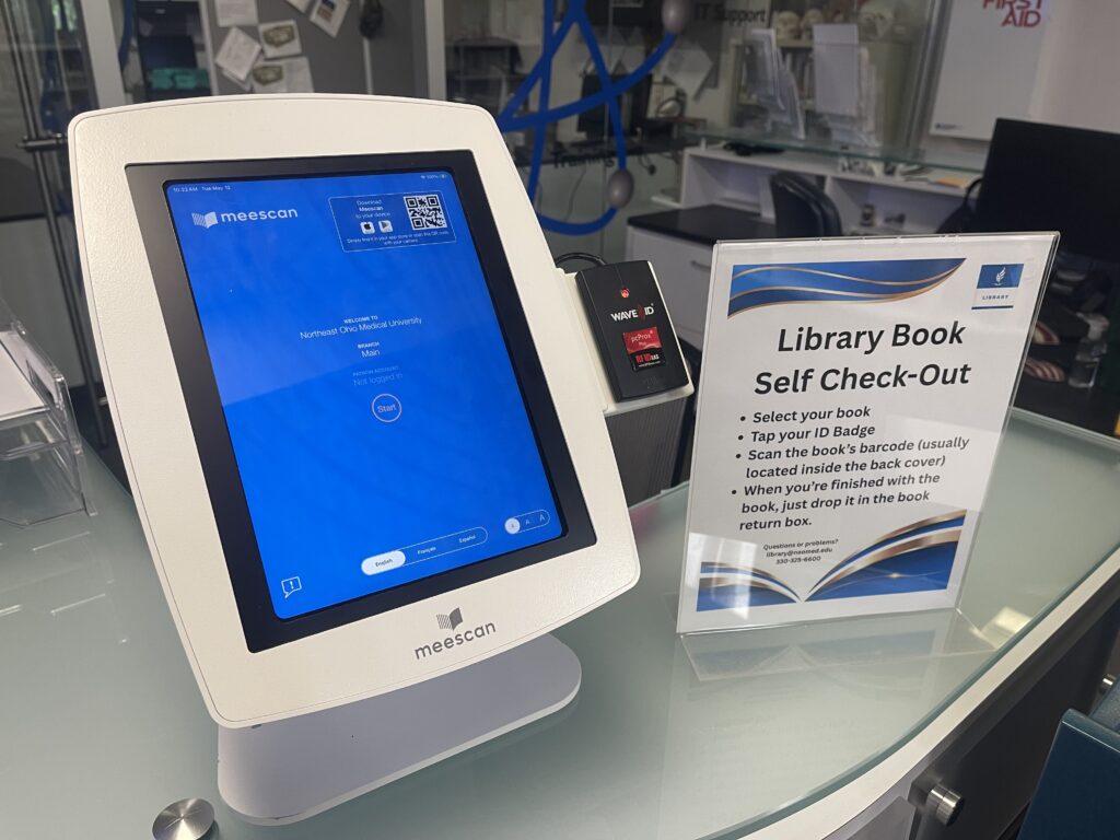 A blue screen device sits next to a sign reading "Library Book Self Check-Out" with additional instructions for using the device
