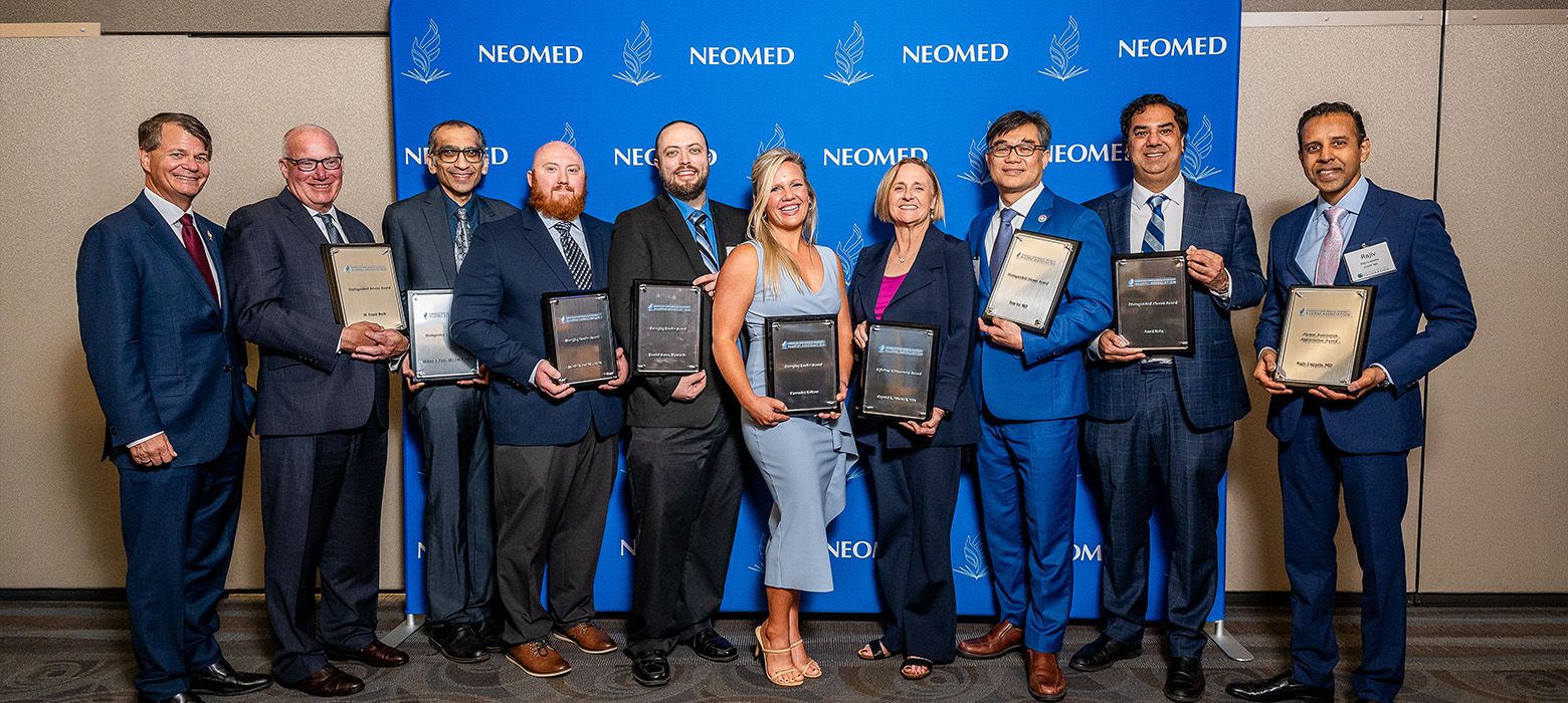 A group of people in business attire holding award plaques pose in front of a NEOMED blue backdrop.