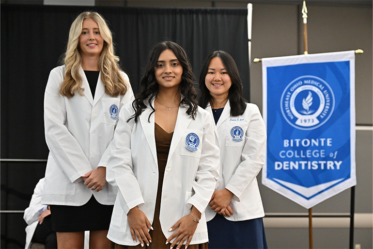 A new student receives her white coat from two other students in white coats. The gonfalon of the Bitonte College of Dentistry appears behind them