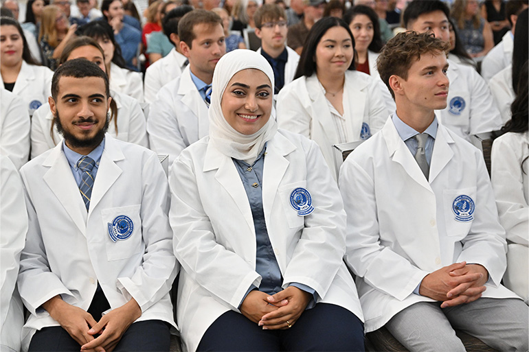 Three students in white coats sit side by side in an assembly hall