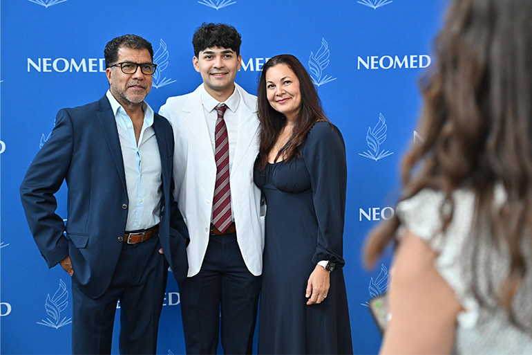 A young man in white coat poses with his parents in front of a NEOMED backdrop