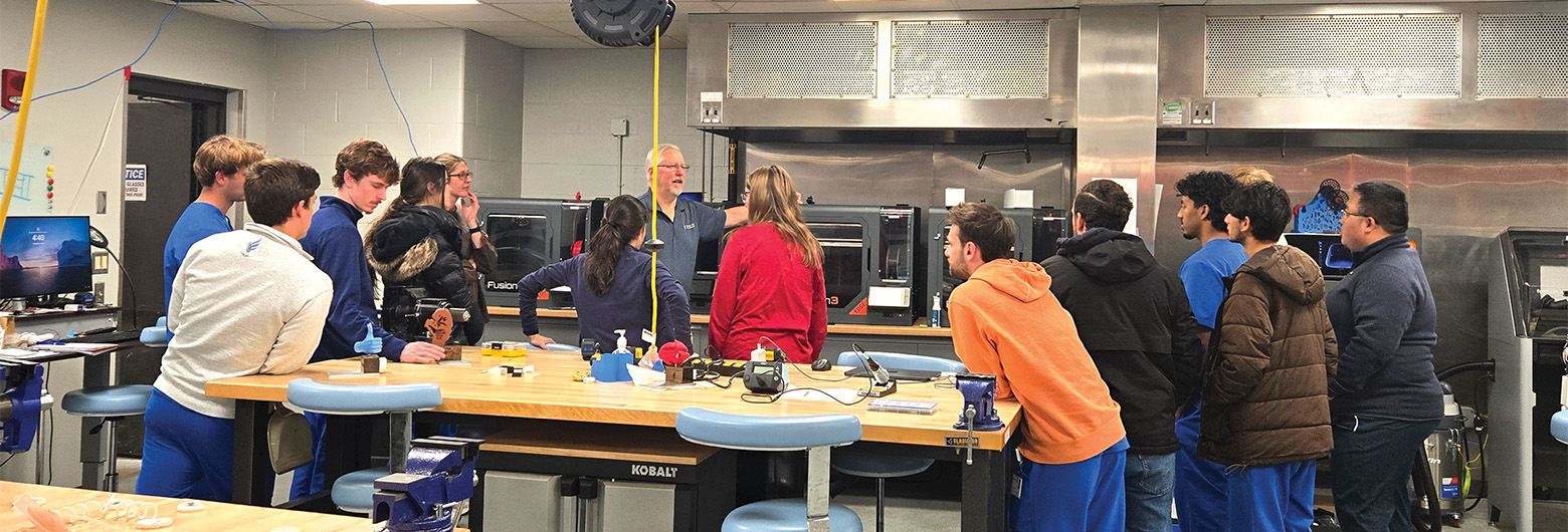A group of students gatherr around a workshop bench to listern to the instructor in the center