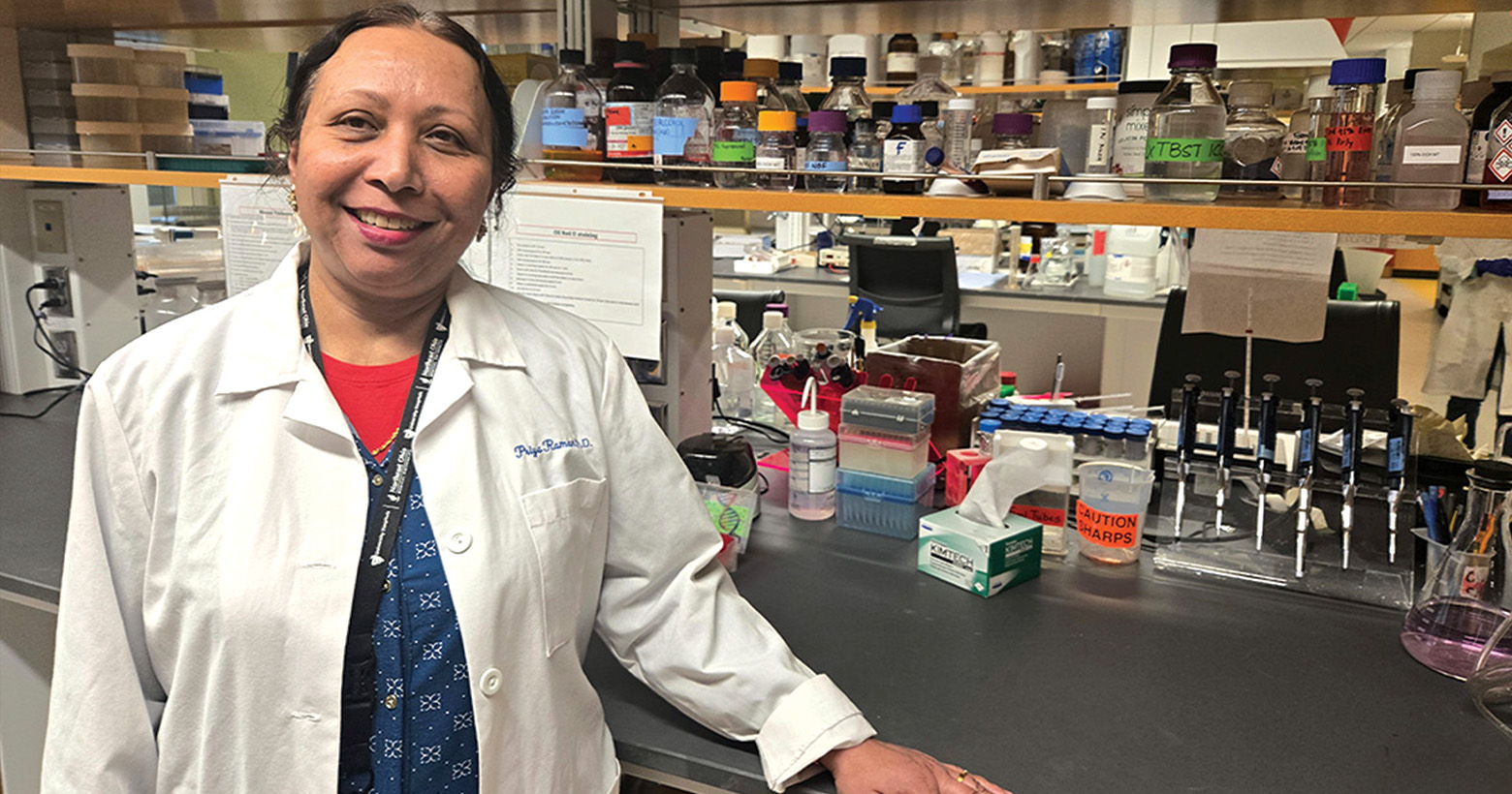 A woman in a lab coat rests her arm on a lab bench