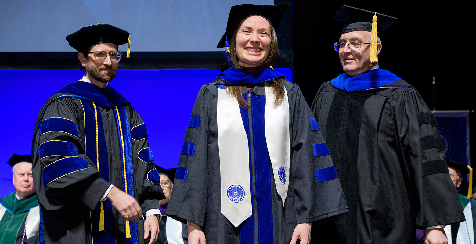 A smiling woman receives her Ph.D. hood from two faculty members