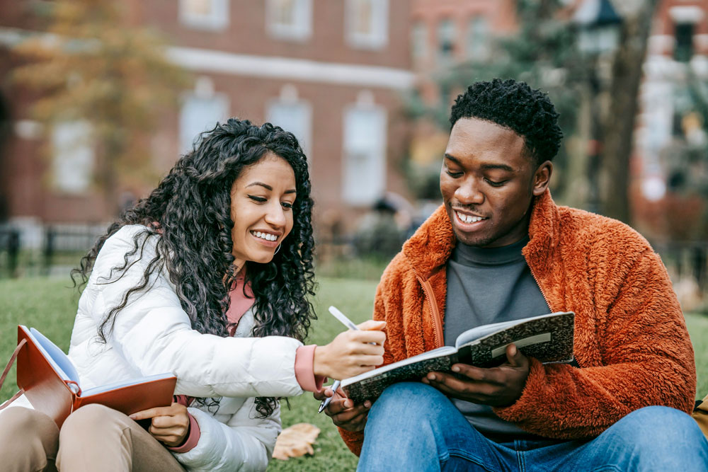Two students sit on a campus lawn and smile while writing in an open notebook.