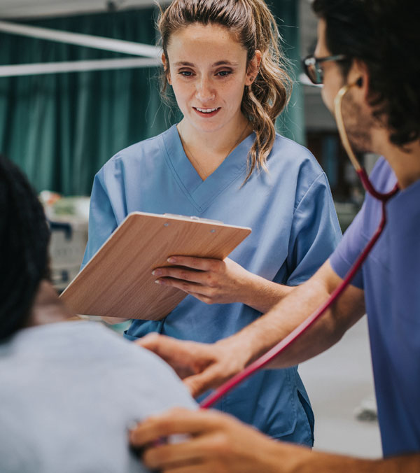 A healthcare professional in blue scrubs reviews a clipboard while another clinician uses a stethoscope to examine a seated patient.