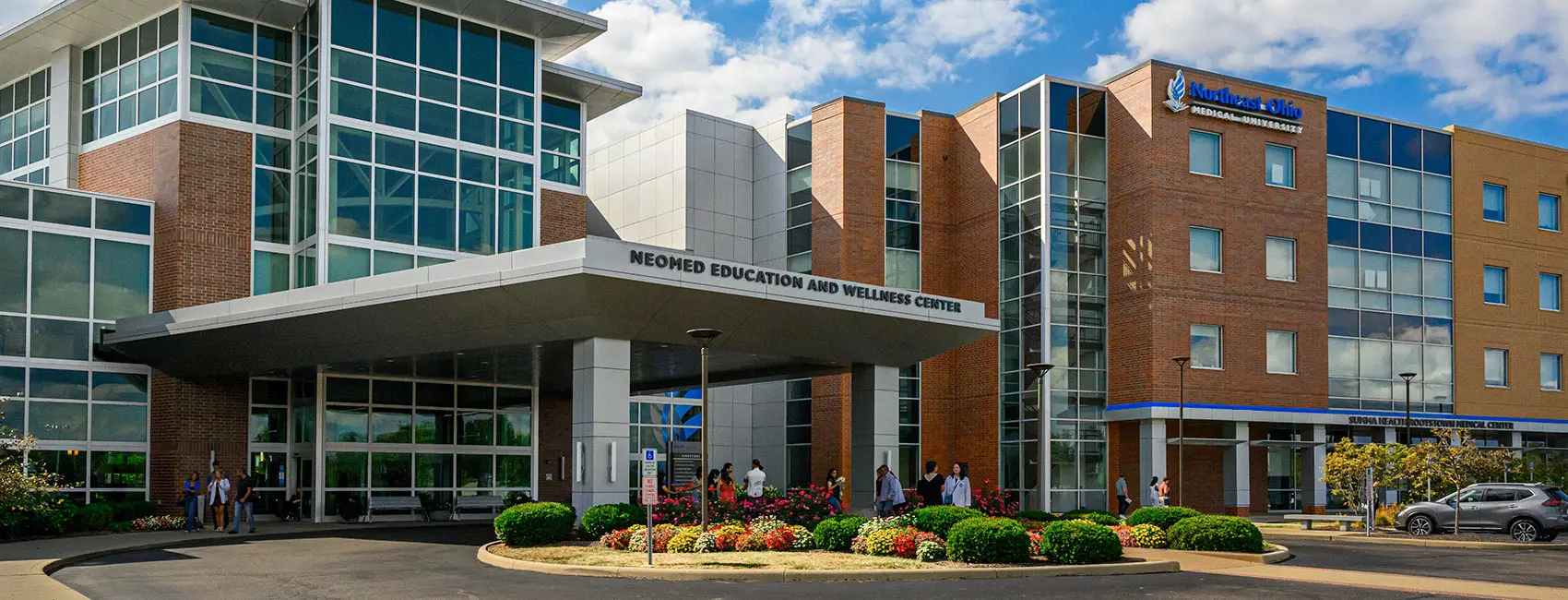 Exterior of the NEOMED Education and Wellness Center, featuring brick and glass buildings, landscaped gardens and people near the main entrance.