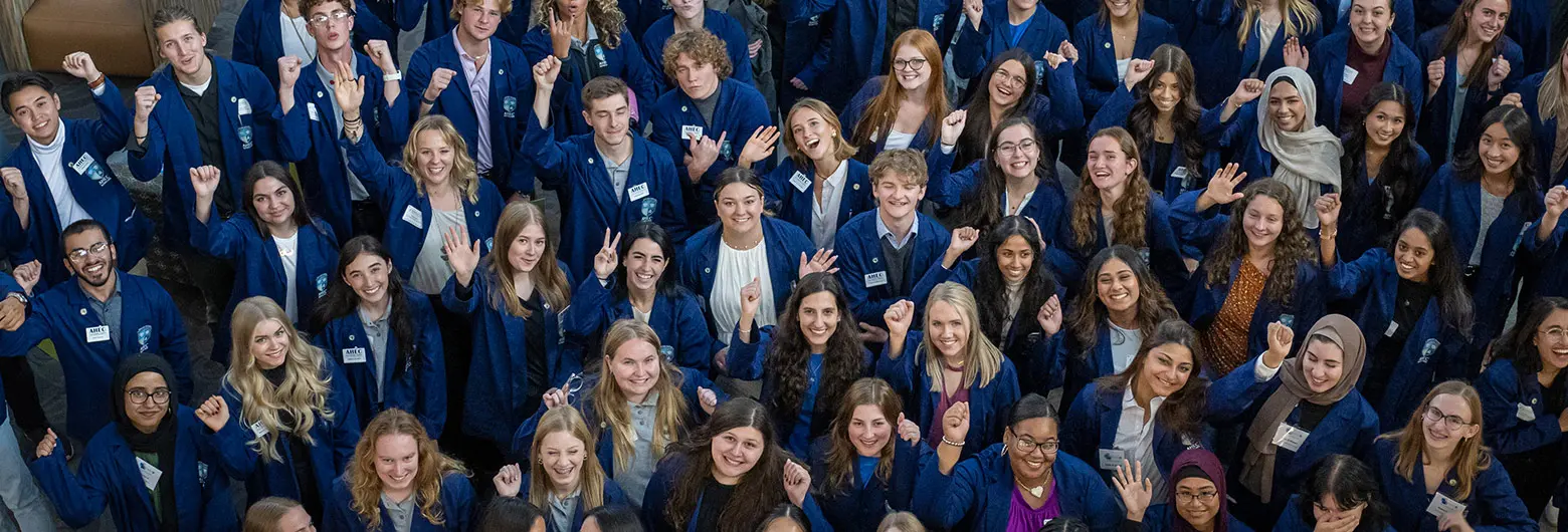 A large, diverse group of AHEC scholars in matching navy jackets smiles, waves and raises their fists for a group photo.