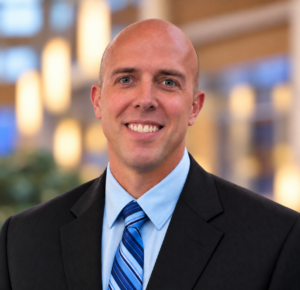 Professional headshot of a smiling bald man in a suit and tie.