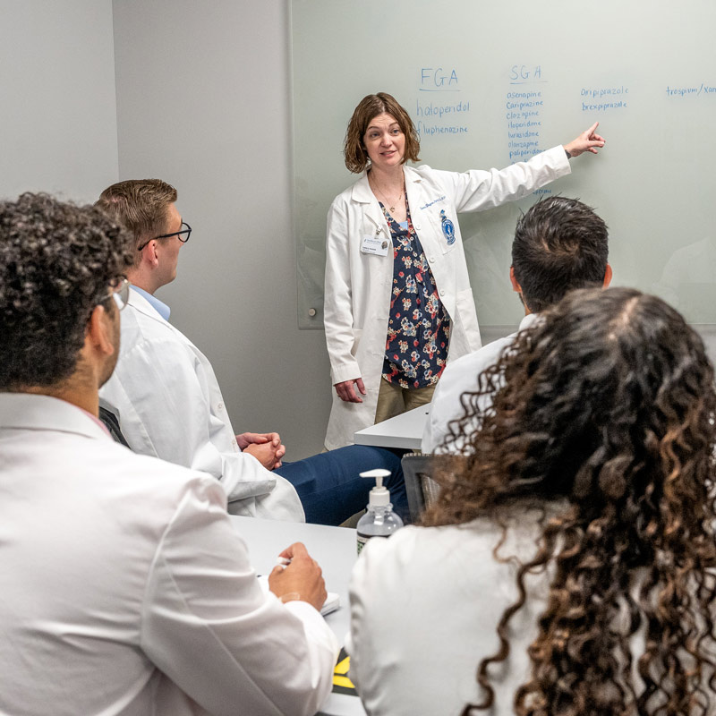 A faculty member in a white coat points to a list of medications on a whiteboard while teaching a small group of medical students.