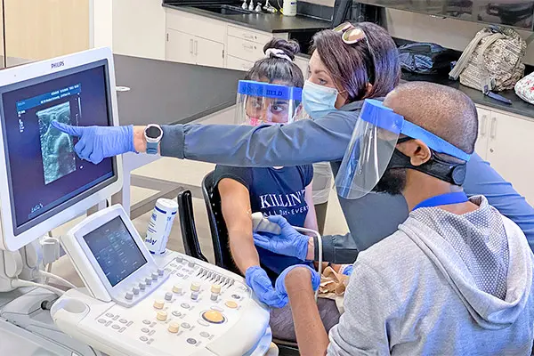 Three people wearing face shields and gloves examine an ultrasound image while one person operates the ultrasound probe on a seated patient’s arm.