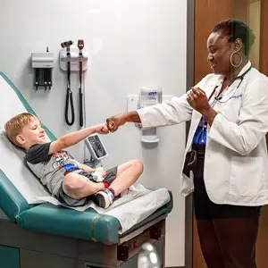 A medical student wearing a stethoscope fist-bumps a young patient seated on an examination table.