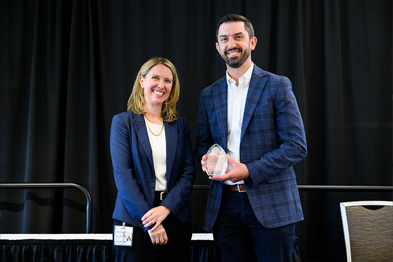 A woman and a man in business attire pose on a stage. The man holds a glass award
