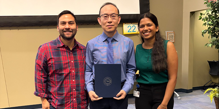 A man in shirt and tie holds an award while posing between a man and a woman