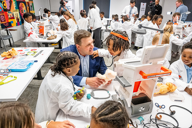 A child in a white coat listens to the heart beat of a man in a sports jacket while another child looks on.