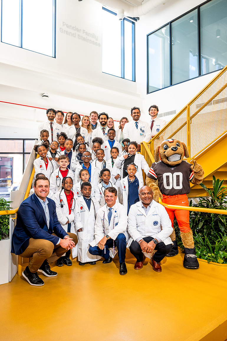 A group of children and adults in white coats pose on as stairway with the Browns mascot and team physician