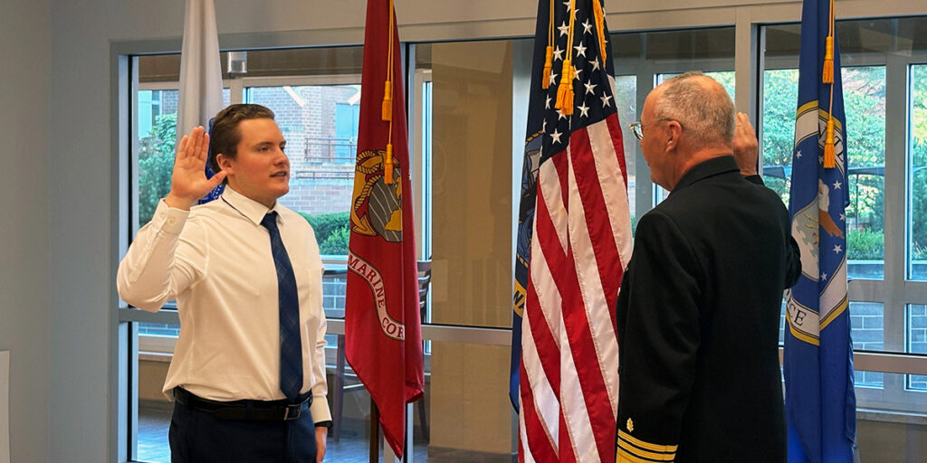 A man in a white dress shirt and tie raises his right hand and recites an oath, administered by a man in a U.S. Navy uniform