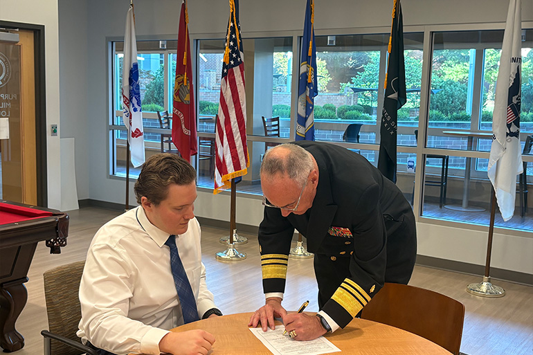 A man in a white dress shirt and tie is seated at a round table. He is watching a man in a Navy uniform sign a piece of paper