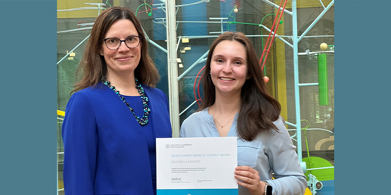 two woman pose in front of a colorful background. The woman on the right holds an award certificate