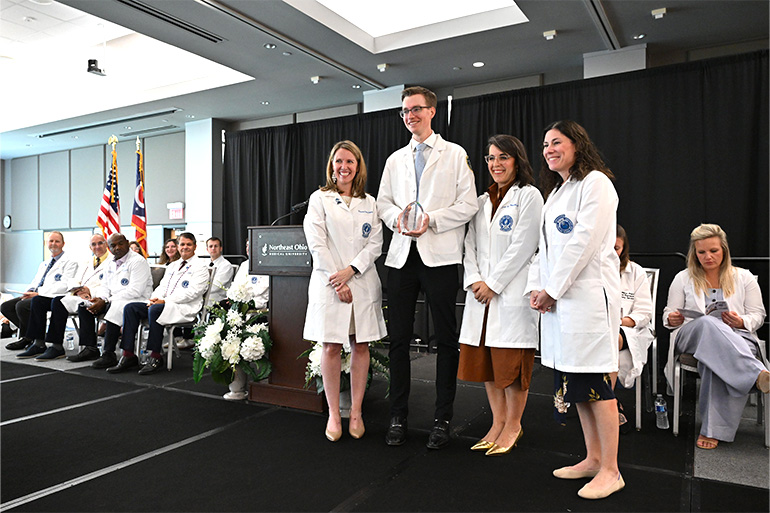 A man and three women, all wearing white coats, pose on a stage