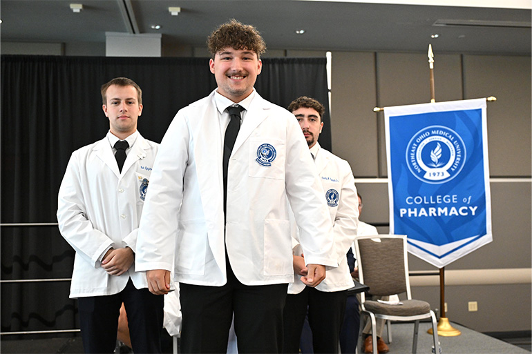 A young man poses after receiving his white coat from two other  students in white coats. The gonfalon of the NEOMED College of Pharmacy appears in the background