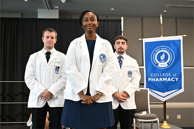 A young woman poses after receiving her white coat from two other students in white coats. The gonfalon of the NEOMED College of Pharmacy appears in the background