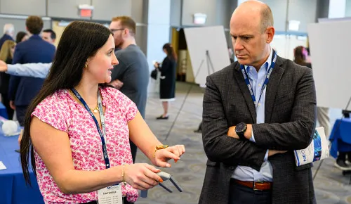 A woman gestures while speaking with a man wearing a conference badge during a professional event, with other attendees and display boards in the background.