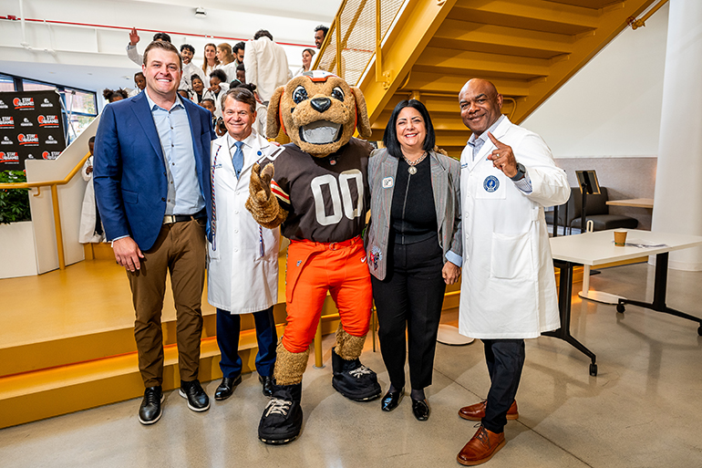 A man in a sports jacket, a woman in business attire and two men in white coats pose with CHOMPS, the Browns mascot, at the bottom of a staircase