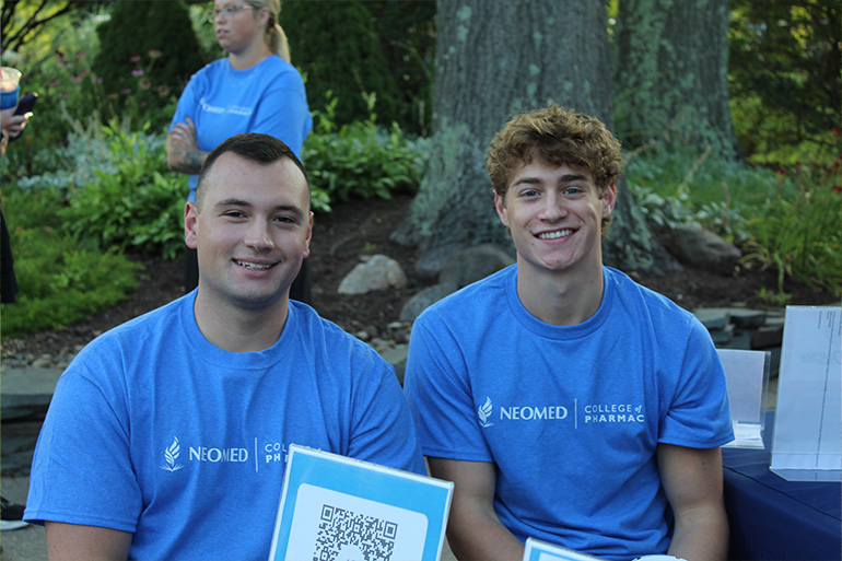two men seated outside. Both are wearing blue NEOMED College of Pharmacy T-shirts