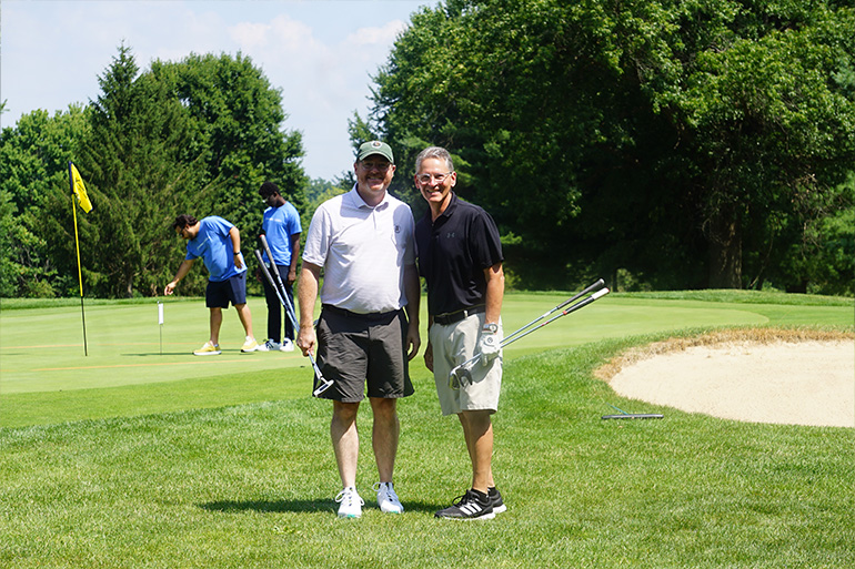 Two men in shorts pose on a golf green, near a sand pit