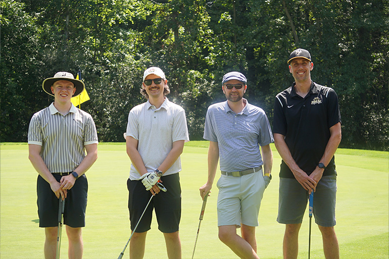 Four men in shorts pose on a golf course. Each man holds a golf club