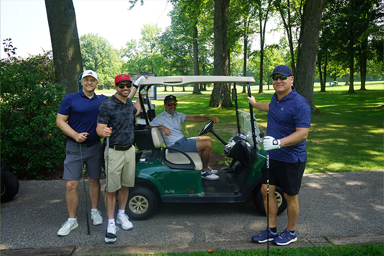 One man sits in a golf cart while three other man pose around it