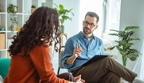 A certified mental health assistant holding a clipboard gestures while speaking with a seated client in a bright office.