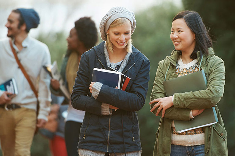 Two female students in light coats chat while walking on a campus.