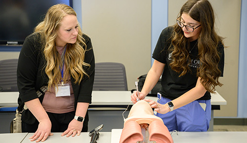 A student in scrubs demonstrates a medical procedure on a pelvic training model while another woman observes in a classroom.