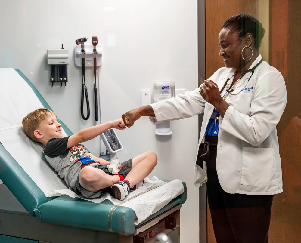 A medical student wearing a stethoscope fist-bumps a young patient seated on an examination table.