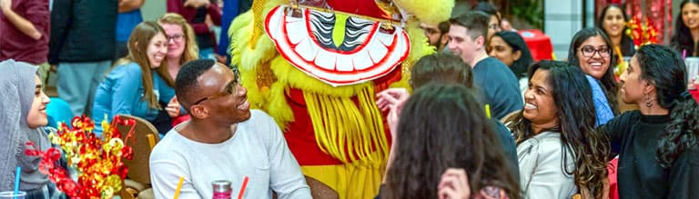 A group of students gather around a costumed character during a campus celebration.