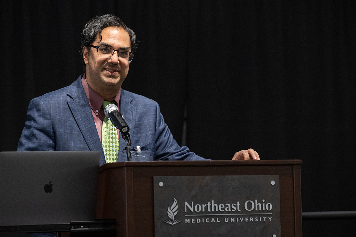 Amol Soin, a man standing at a NEOMED branded podium speaking near a microphone with a laptop on the side in a dark lit room